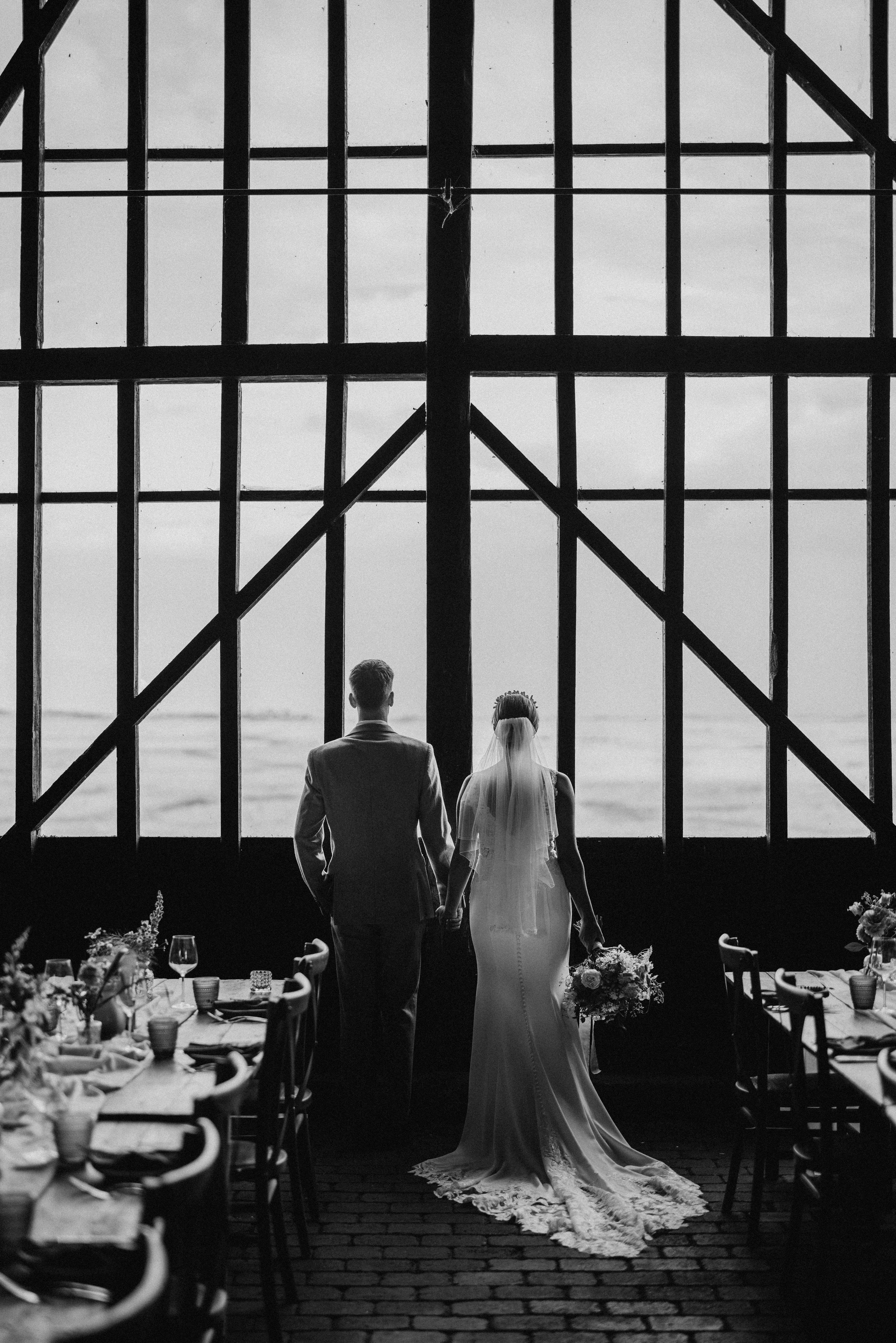A bride and groom, hand-in-hand, looking out of the large glass window in the Elmley Nature Reserve barn. The photo is striking in black and white