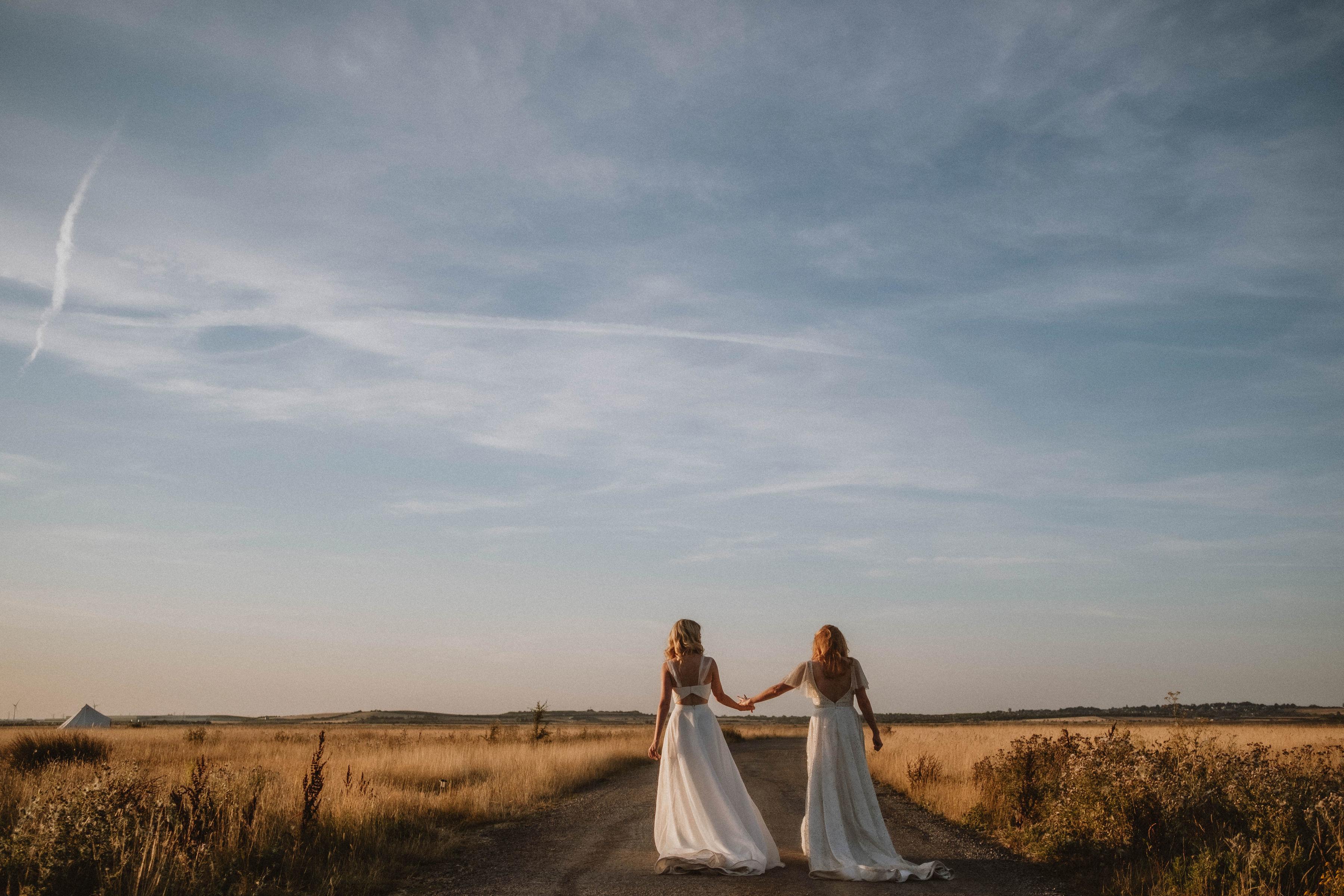 Two brides walk hand-in-hand, backs facing us as they walk through Elmley Nature reserve. The sun is setting, leaving a warm glow across the dried grass fields